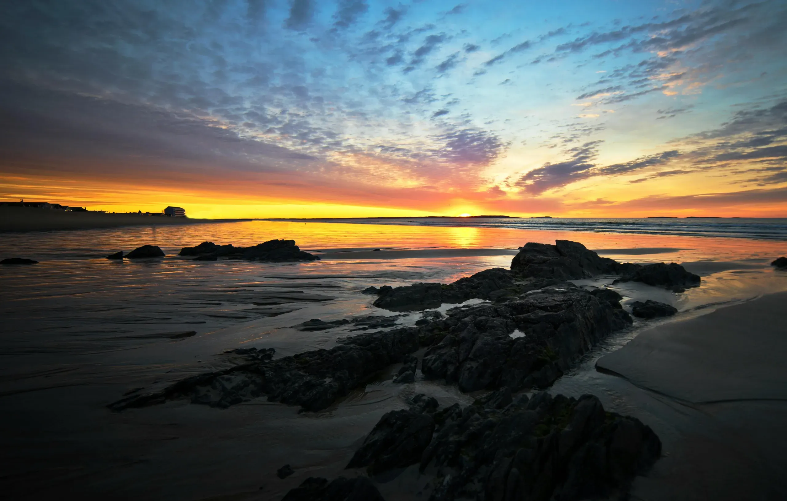 The vibrant colours of a cloudy sunrise are reflected on the wet sand and around the dark rock formations of a shore at Old Orchard Beach, Maine.