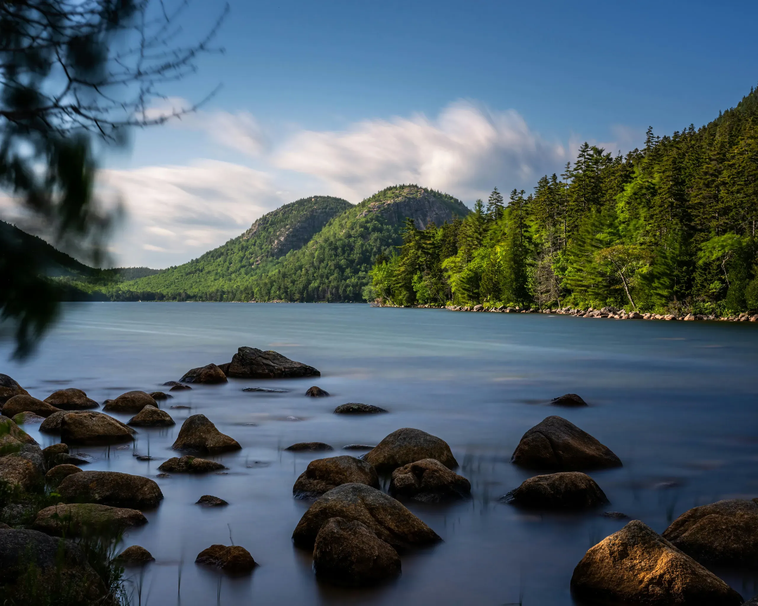 A long-exposure photograph of a river in Maine, showing smooth water with rocks in the foreground and two rouinded, forest-covered mountains in the background.