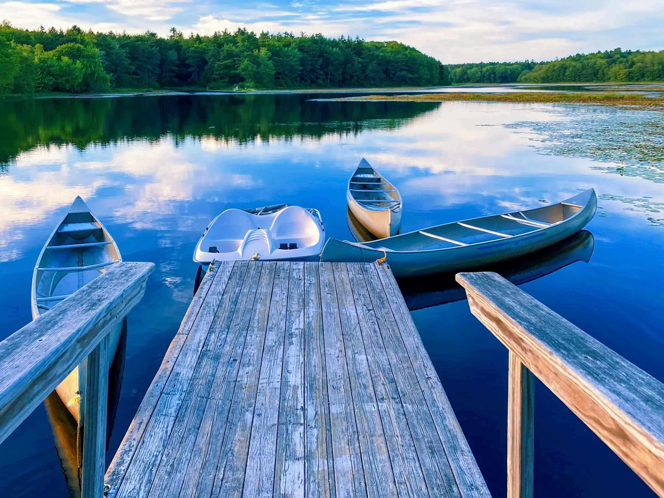 Canoes and a pedal boat are moored at a wooden dock on a calm lake, reflecting the forest and blue sky, in Phippsburg, Maine.