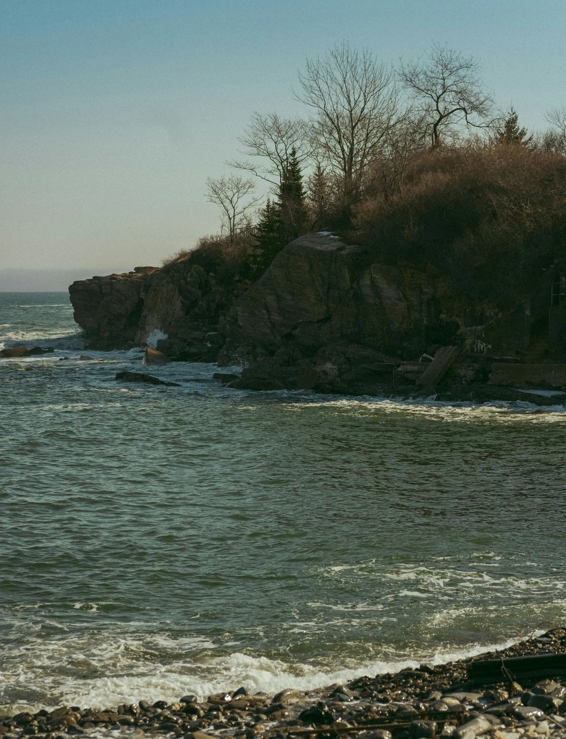A rocky cliff, topped with bare trees and shrubs, juts into the sea in Portland, Maine.