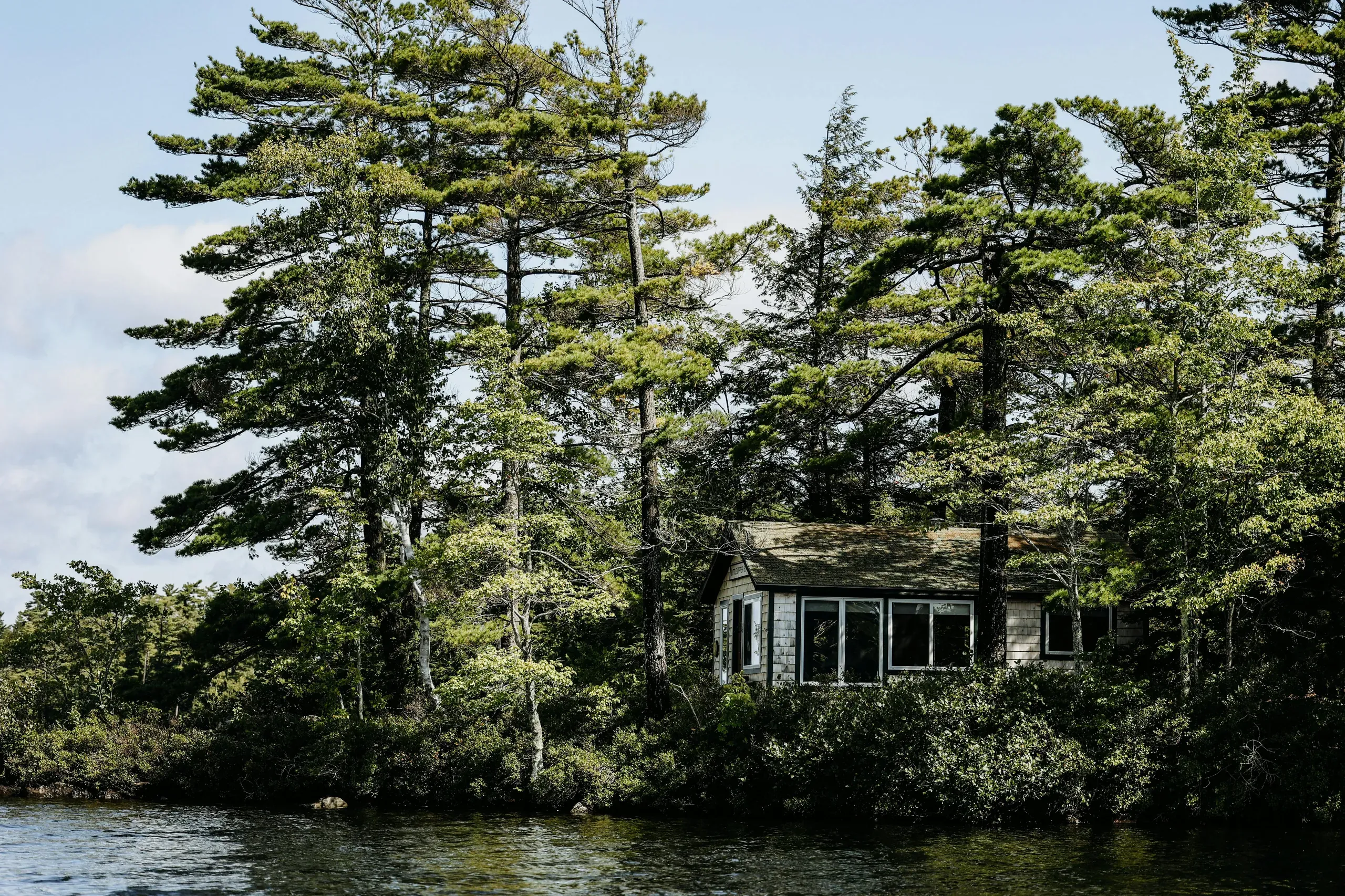 A small white wooden house surrounded by trees sits on the shore beside a body of water in Damariscotta, Maine.