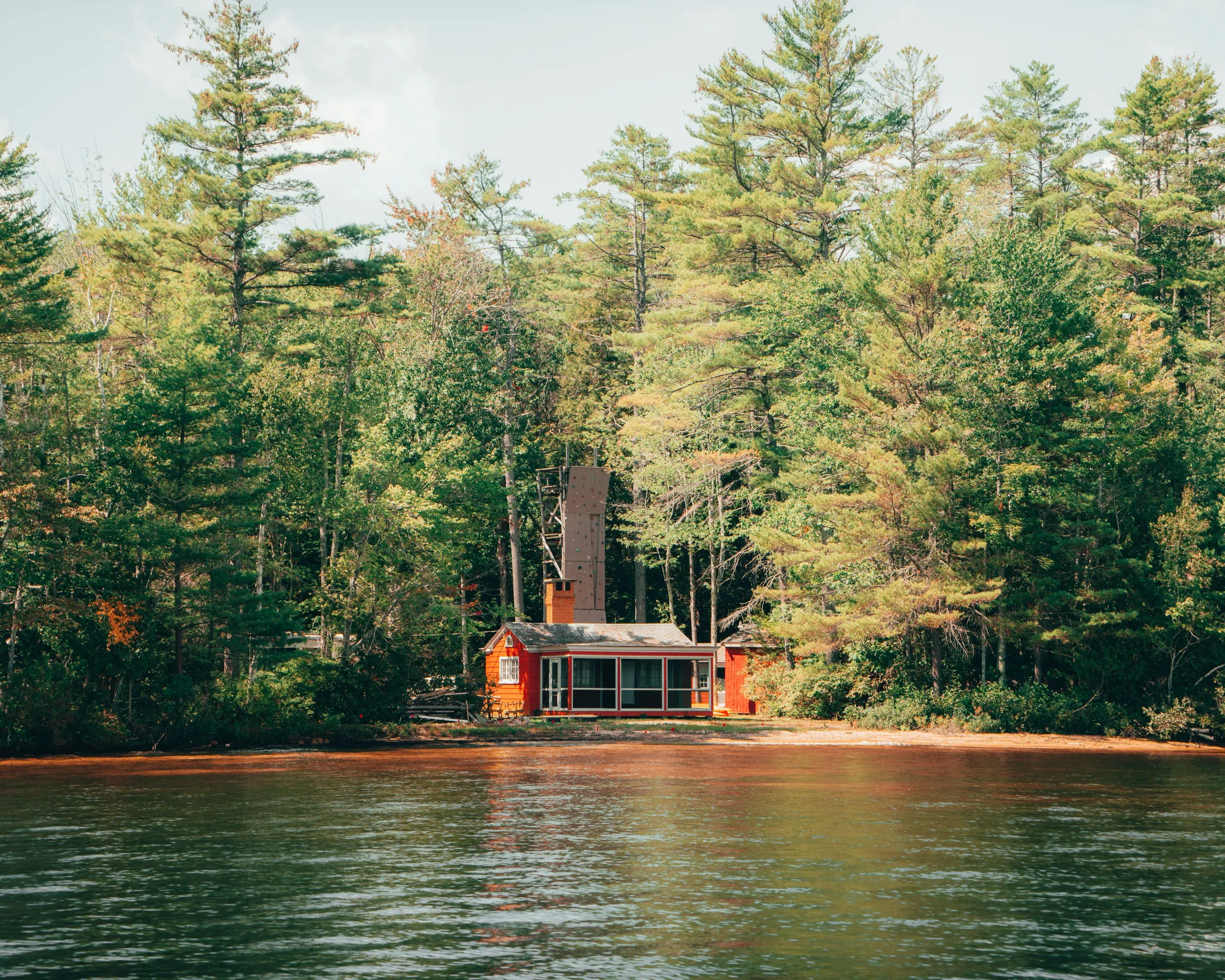 A small, red cabin with a screened-in porch sits on the shore of a lake, at the edge of a dense pine forest in Norway, Maine.