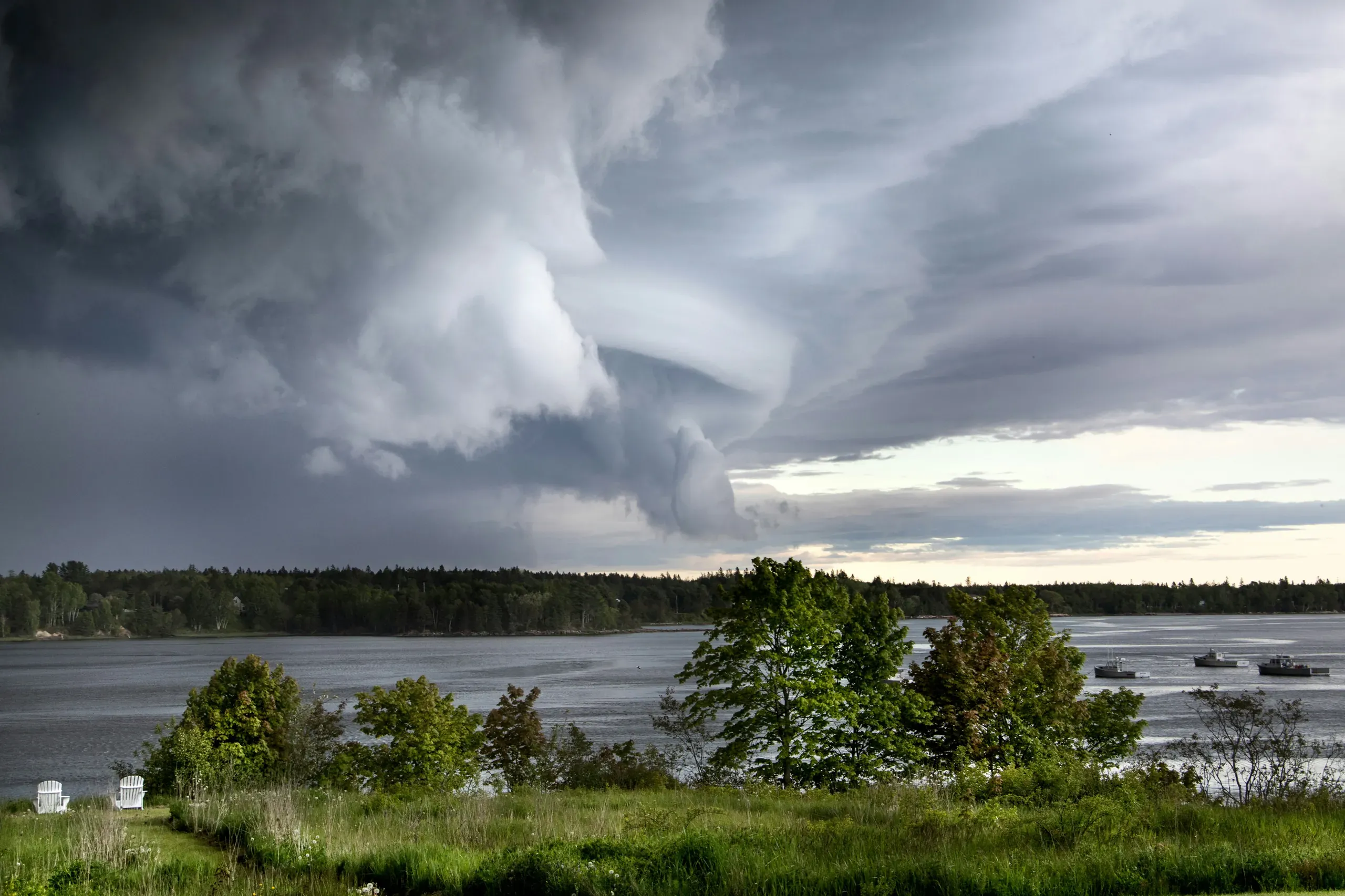 A dramatic, grey storm sky gathers over a body of water, viewed from a grassy shore with two white chairs in Maine.