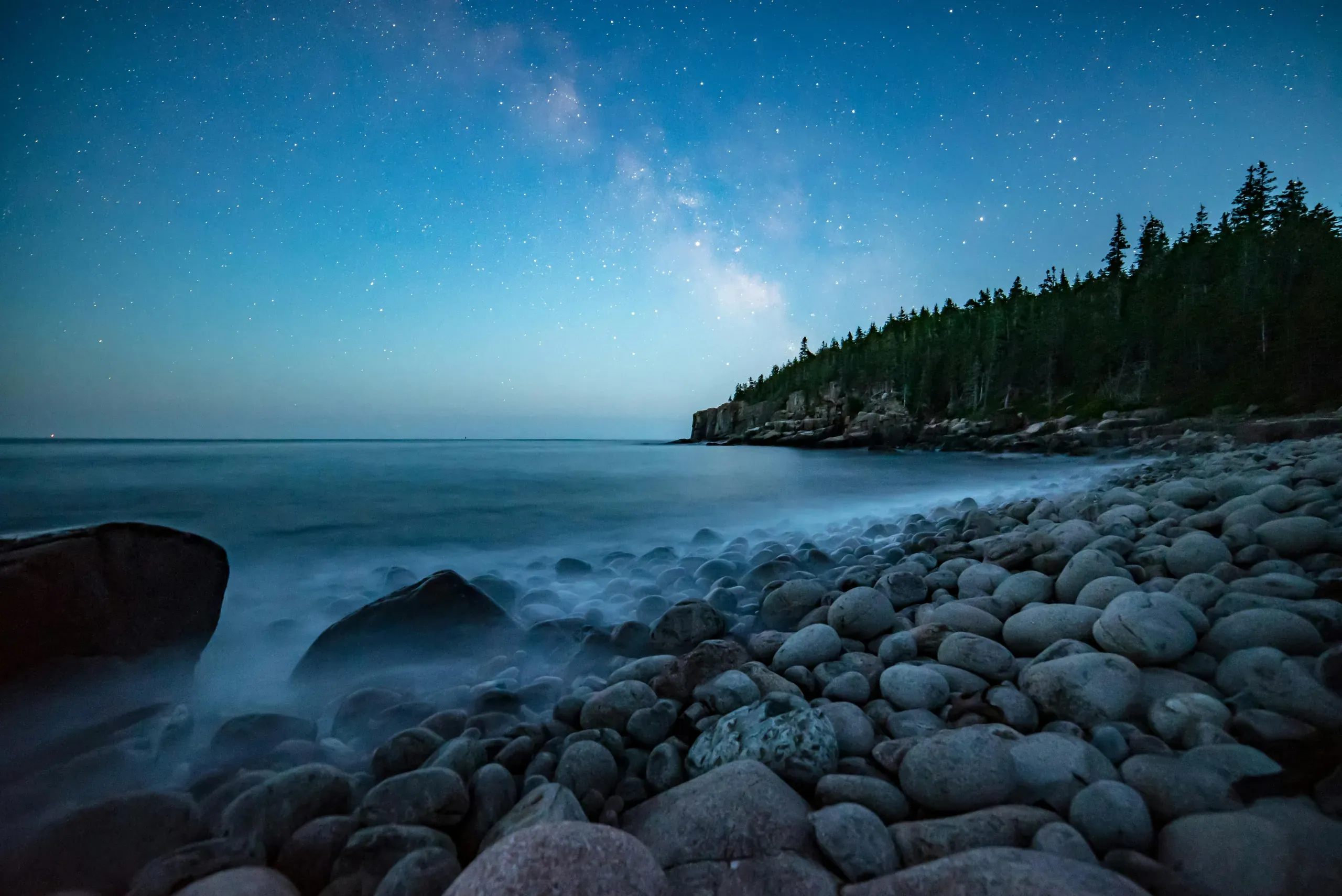 A long-exposure photograph of the rocky shore in Bar Harbor, Maine, where misty water flows over rounded stones under a night sky dense with stars.