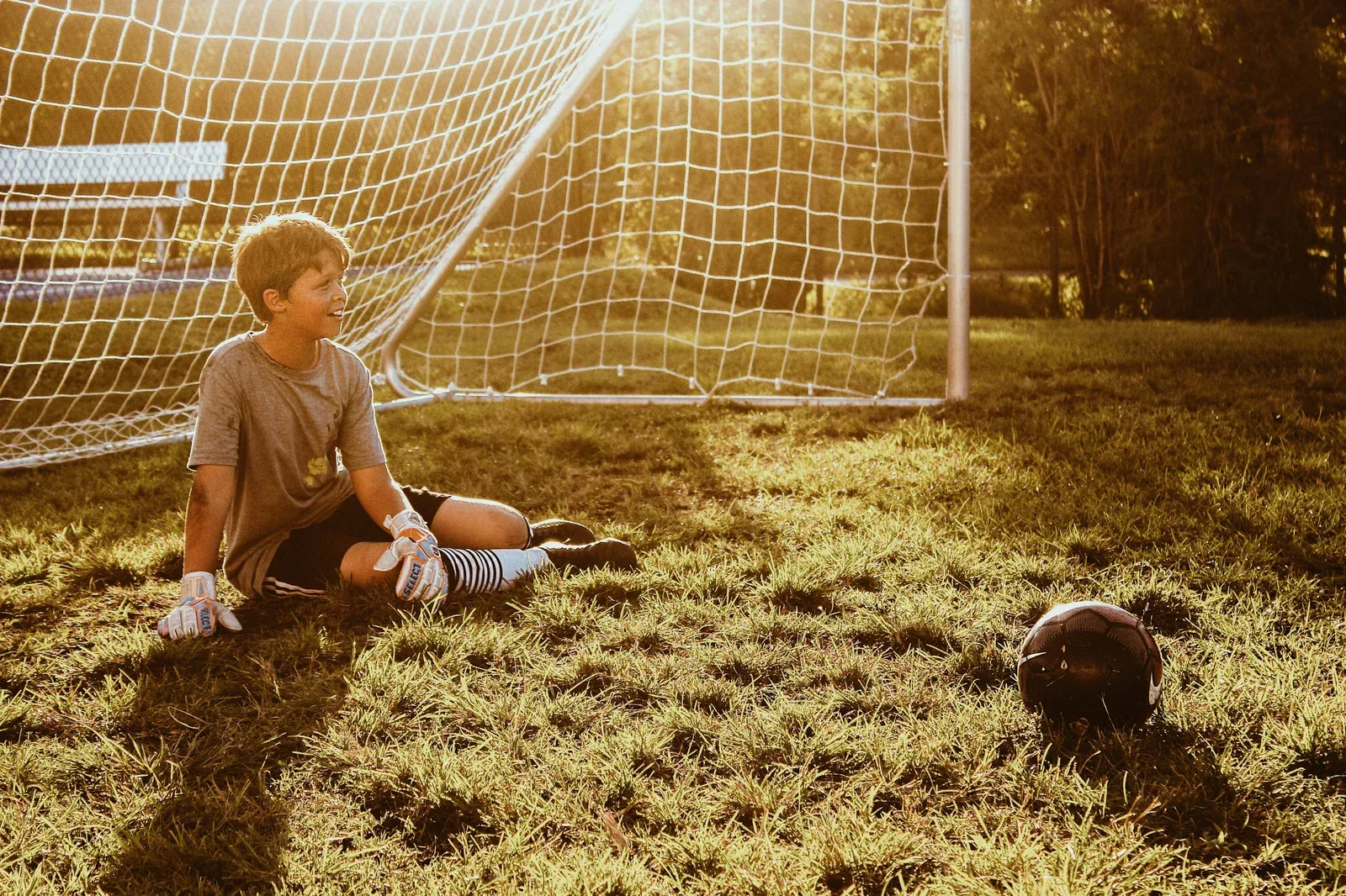 A young boy sitting on the grass in front of a football goal in golden evening sunlight, a ball beside him