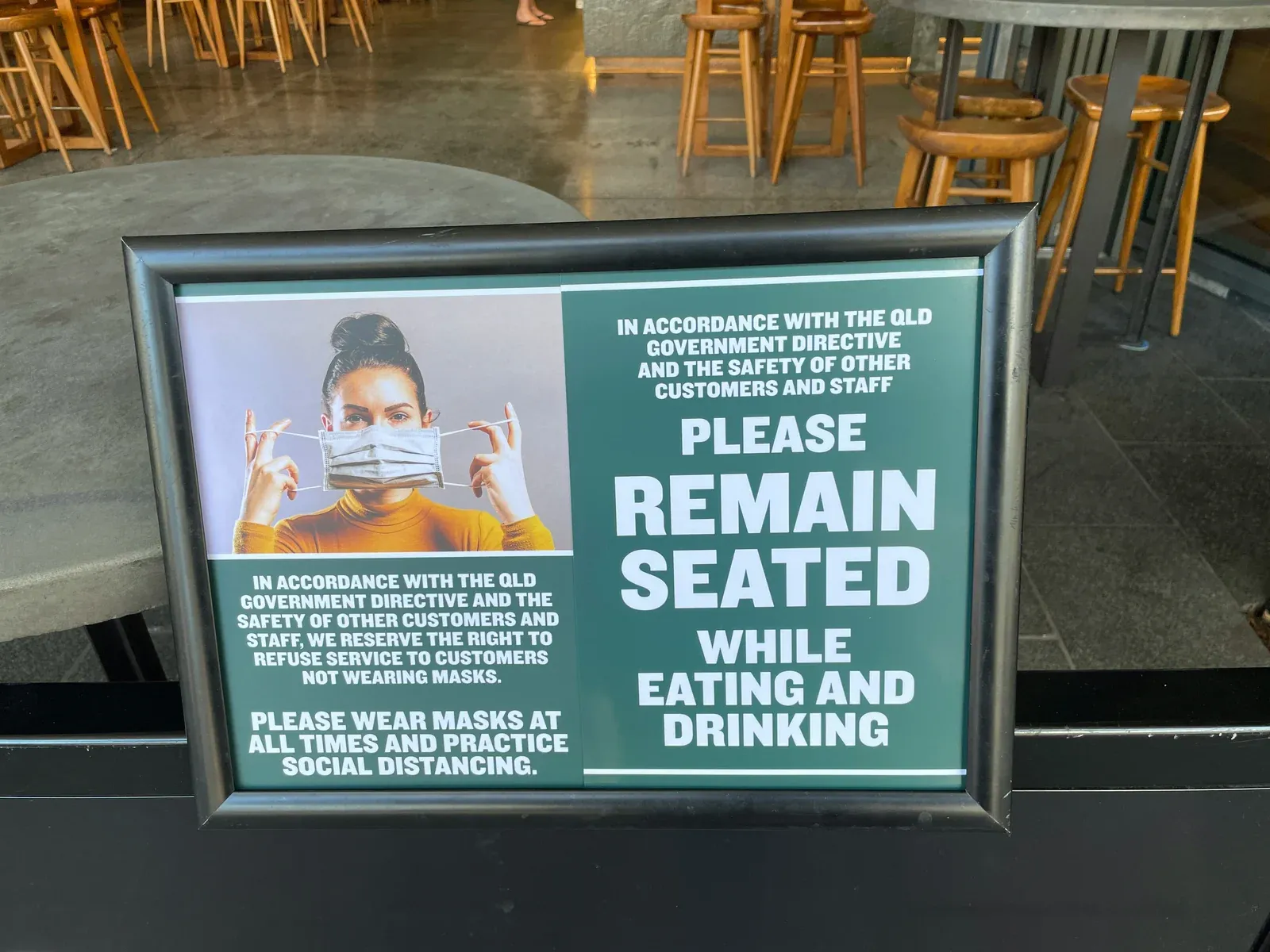 Framed sign in an Australian cafe reading 'Please remain seated while eating and drinking' and 'Please wear masks at all times and practice social distancing', in accordance with the QLD Government directive