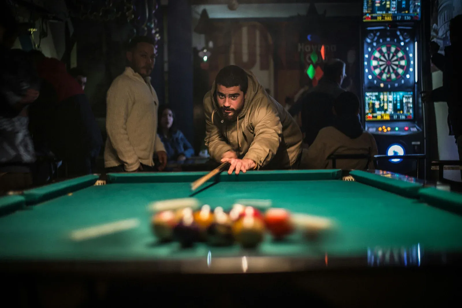 A man lining up a pool shot in a dimly lit bar, racked balls in the foreground and a dartboard glowing behind him
