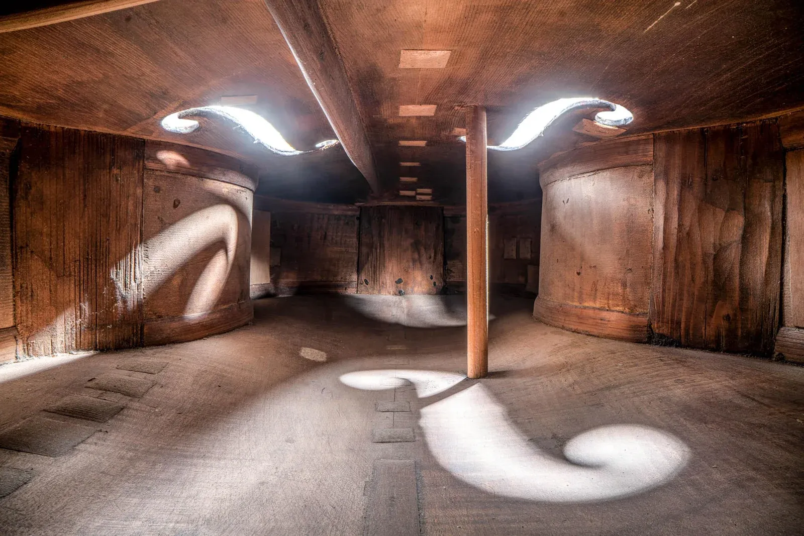Photo taken from inside a 240-year-old cello, showing the wooden interior with the sound post, f-holes casting light, and curved wooden walls