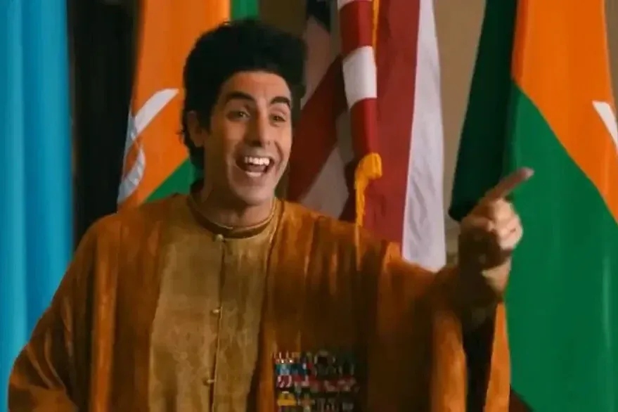 Sacha Baron Cohen as Admiral General Aladeen in The Dictator, laughing in a gold tunic in front of flags