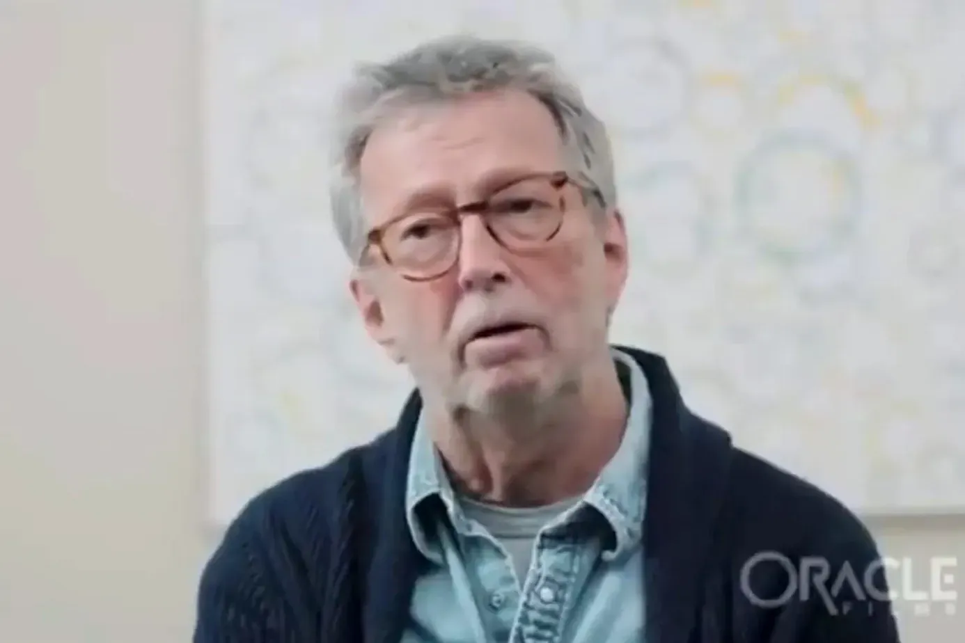 Eric Clapton speaking in an interview, wearing glasses and a dark cardigan over a denim shirt, with an Oracle Films watermark