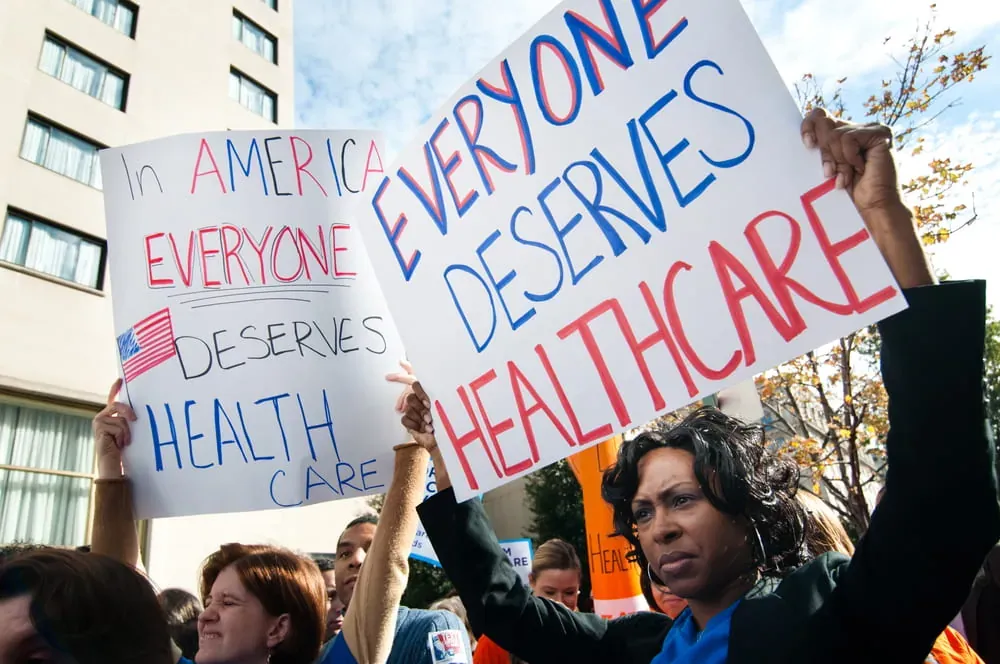 Healthcare protest photo: demonstrators hold handwritten signs reading In America Everyone Deserves Health Care and Everyone Deserves Healthcare outside a building