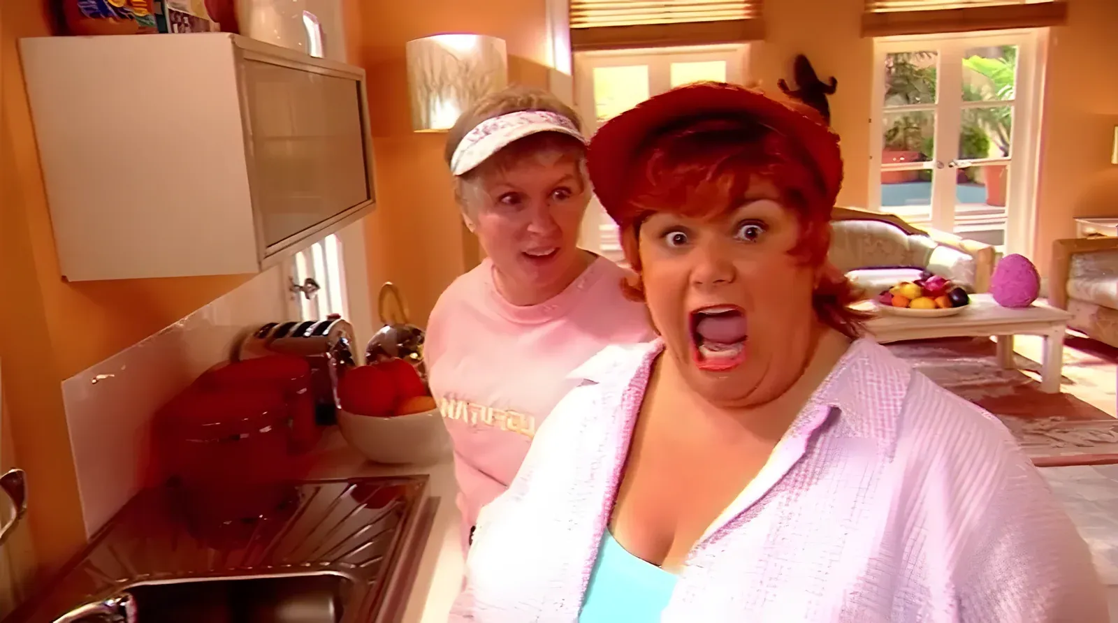 French and Saunders in character in a Florida kitchen - Dawn French with red hair and hat looking shocked at the camera, Jennifer Saunders behind in a visor