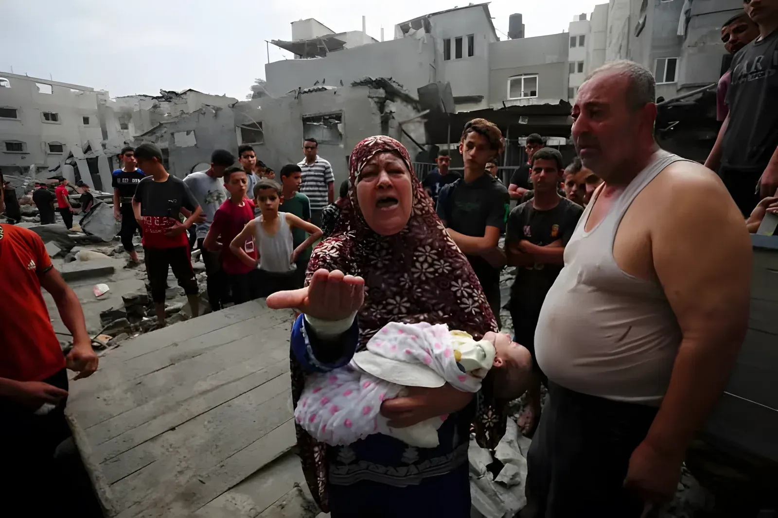 A Palestinian woman crying out in anguish while holding a baby, surrounded by people and destroyed buildings in Gaza