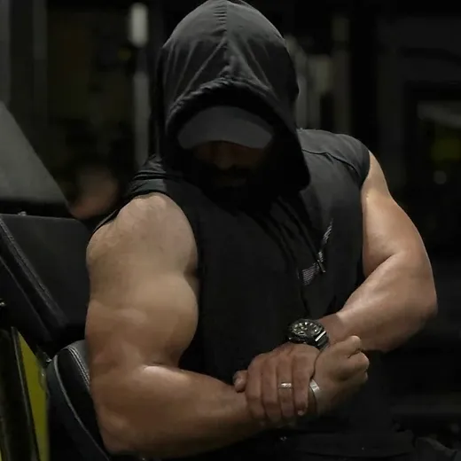 Hasan Almoghani training in a gym, wearing a dark hooded sleeveless top, his massive arms crossed, face hidden in shadow