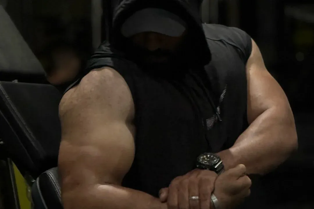 Hasan Almoghani training in a gym, wearing a dark hooded sleeveless top, his massive arms crossed, face hidden in shadow