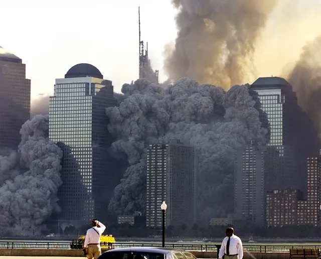 View from the New Jersey waterfront with two people in the foreground, massive dust cloud billowing behind World Financial Center towers after WTC collapse, antenna mast visible through the smoke