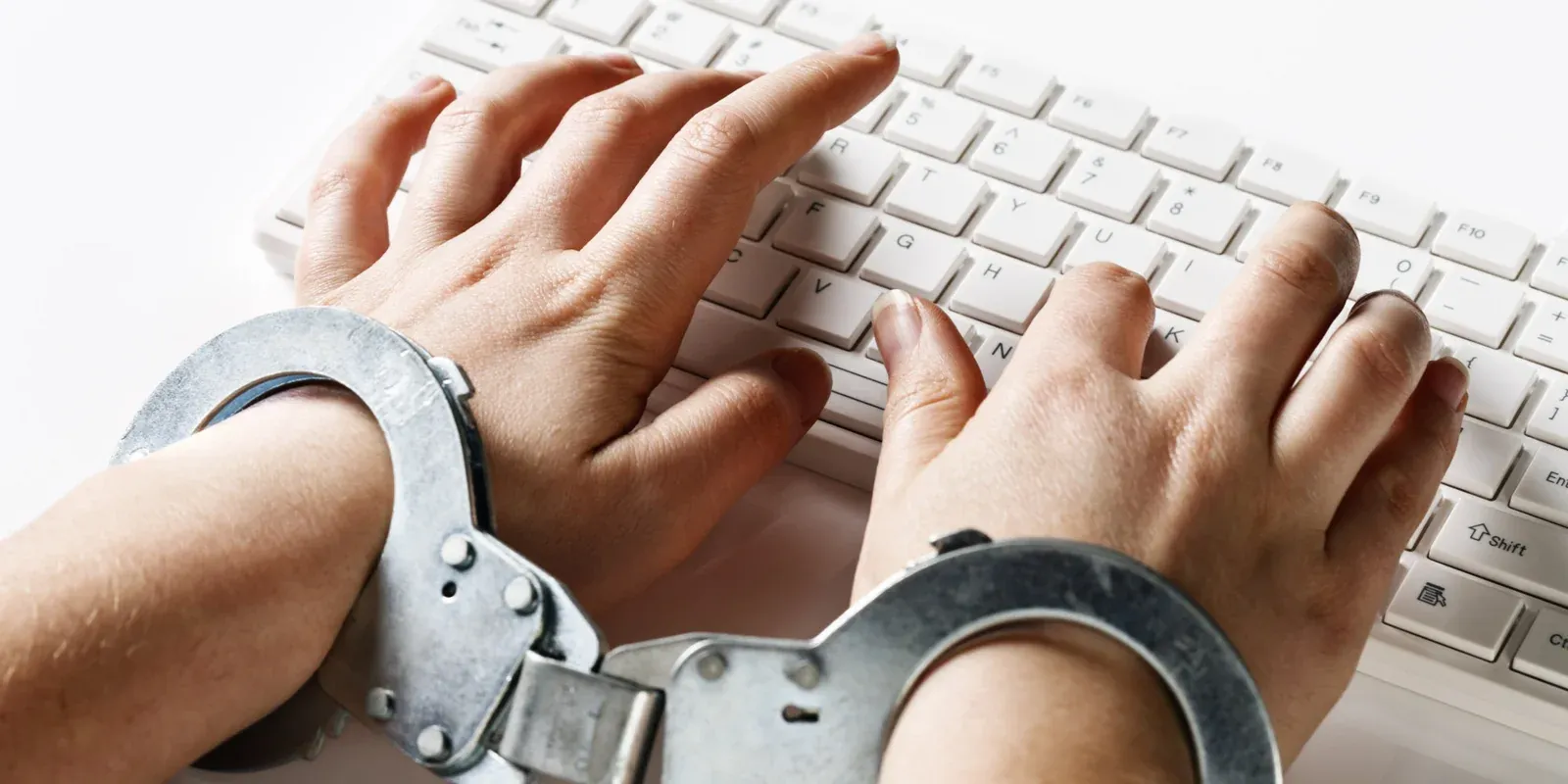 Hands in steel handcuffs typing on a white computer keyboard