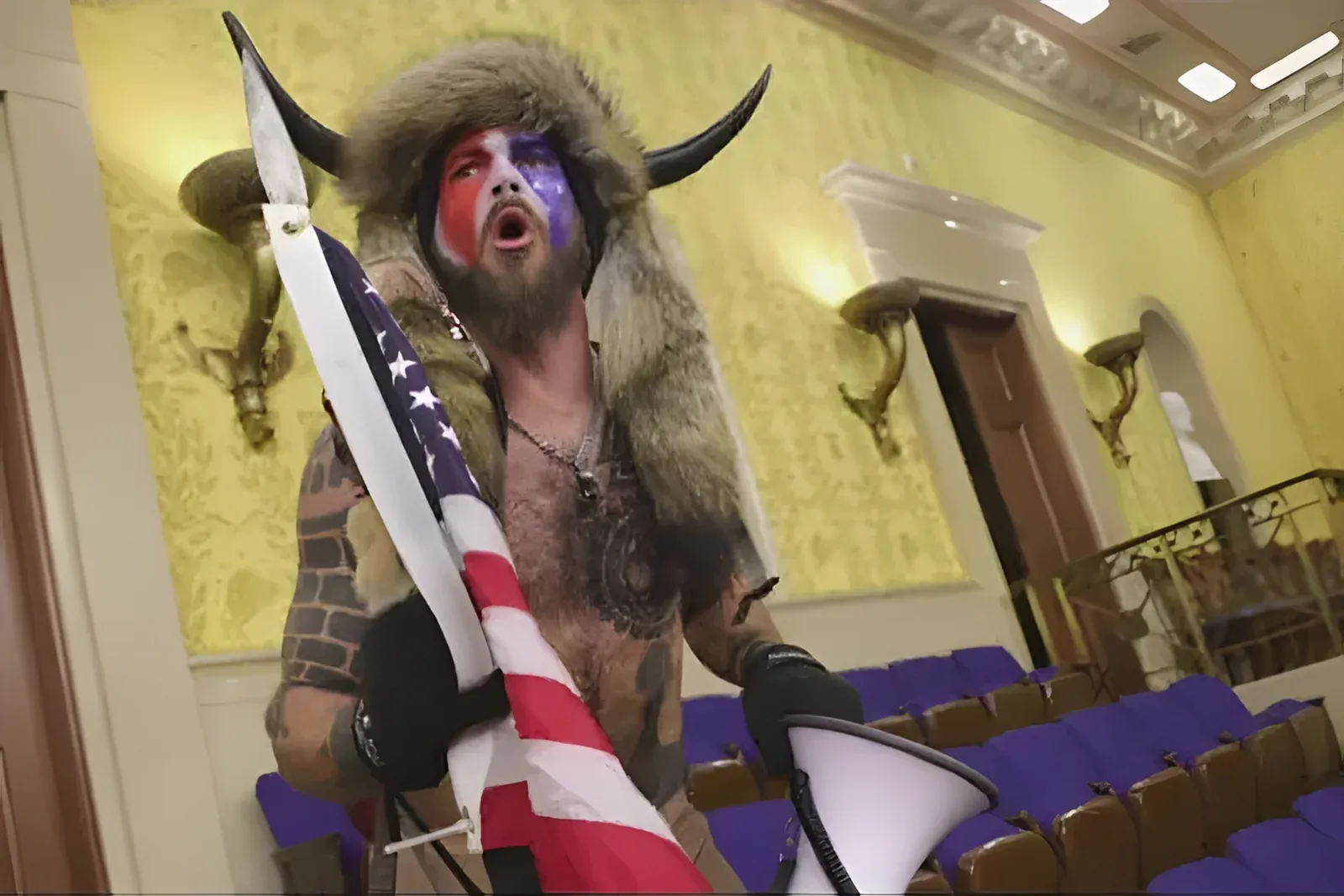 Jacob Chansley inside the US Capitol on January 6th, shirtless with face paint, fur hat with horns, carrying an American flag and a megaphone