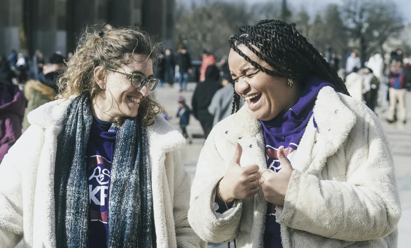 Two women laughing together outdoors, one in glasses and a scarf, the other in a cream fleece jacket