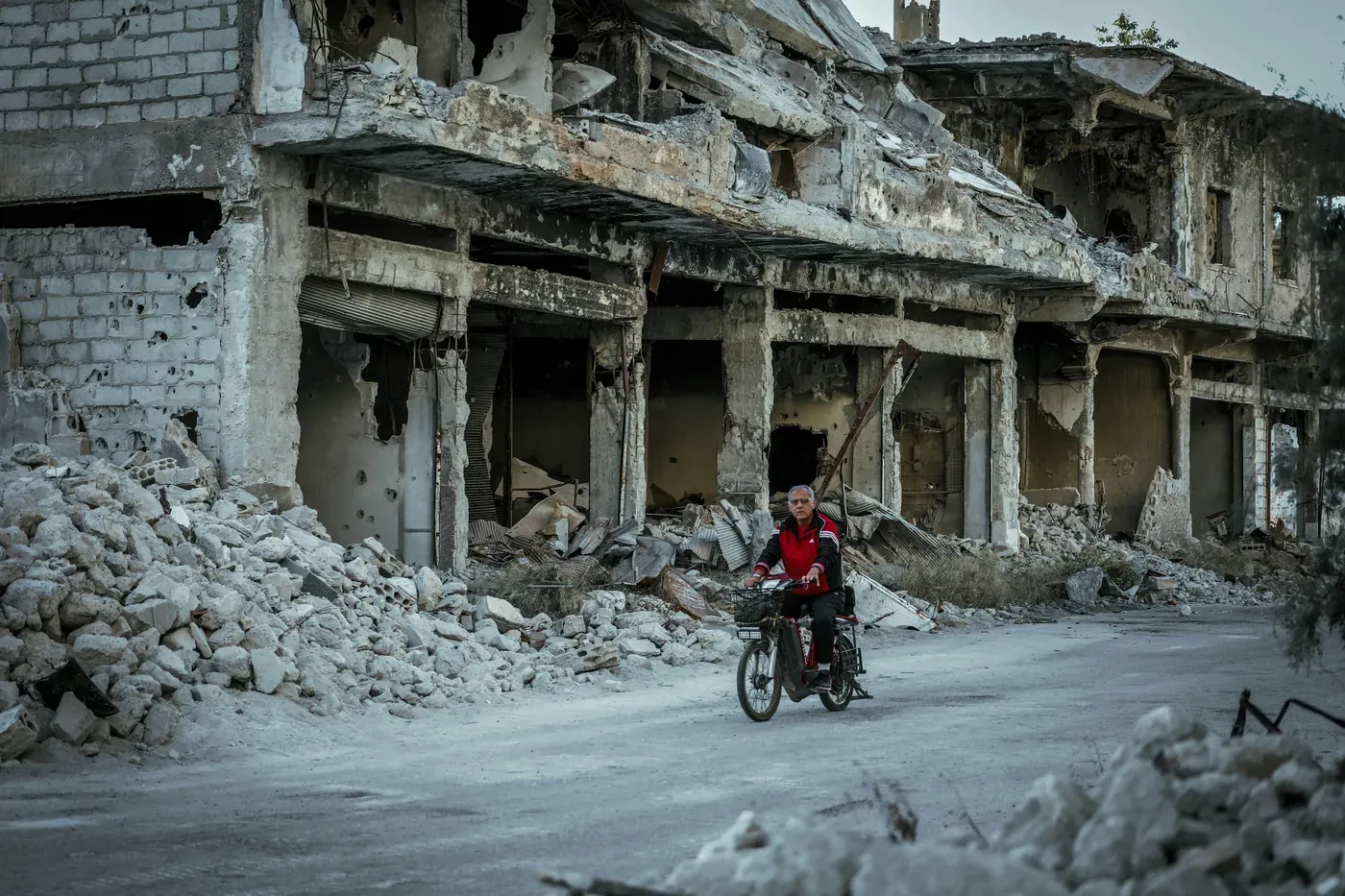A boy cycling past a row of bombed-out buildings, rubble spilling onto the road