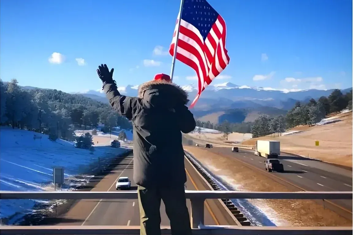 Man standing on a highway overpass holding a large American flag and waving to vehicles passing below, with snow-dusted mountains in the background