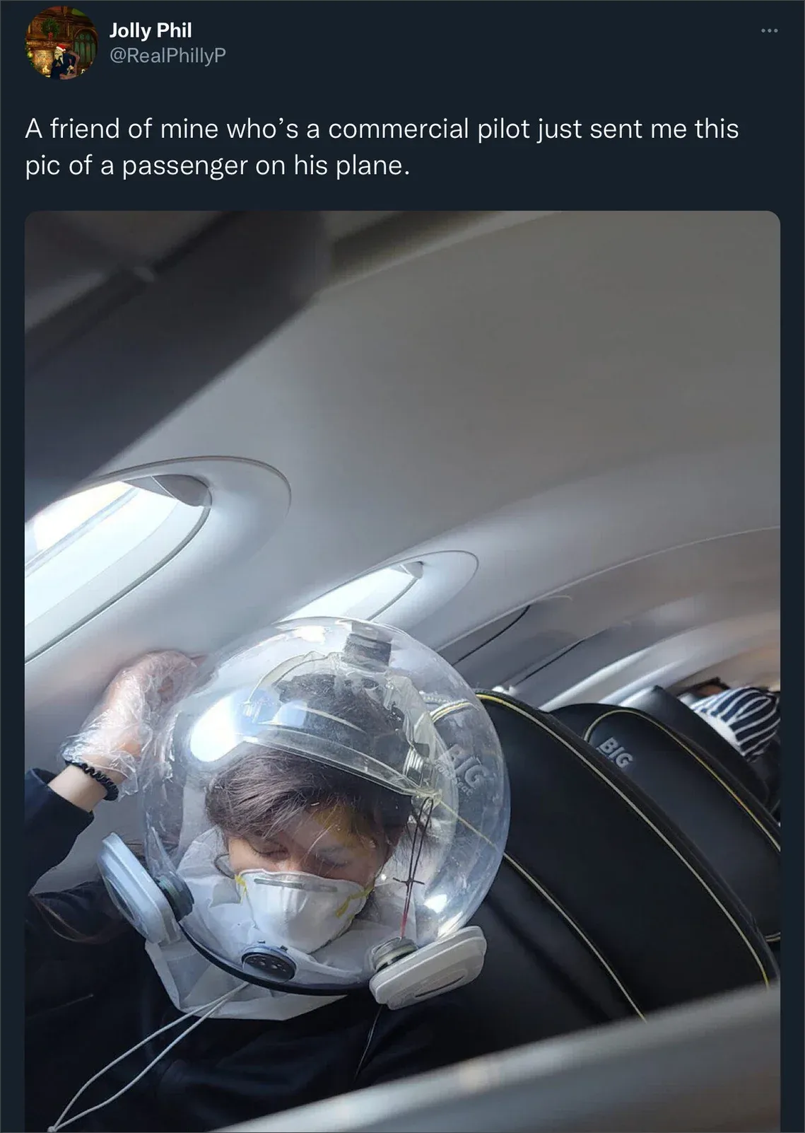 Tweet from @RealPhillyP showing airline passenger wearing clear plastic bubble helmet over their head with mask underneath