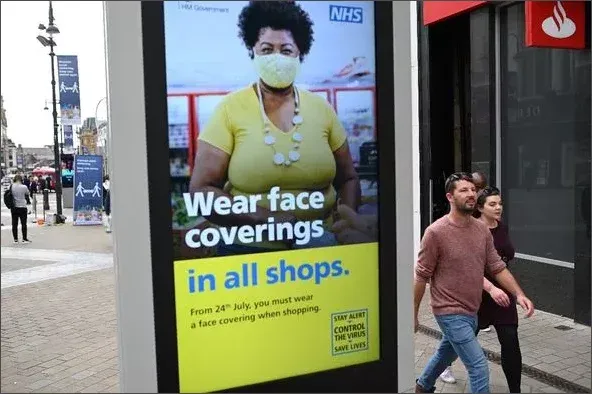 UK Government and NHS public health poster reading Wear face coverings in all shops from 24th July, with two unmasked pedestrians walking past the sign on a high street