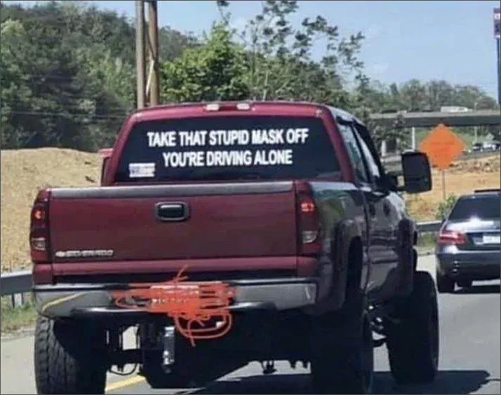 Red pickup truck with large white text on rear window reading TAKE THAT STUPID MASK OFF YOU'RE DRIVING ALONE