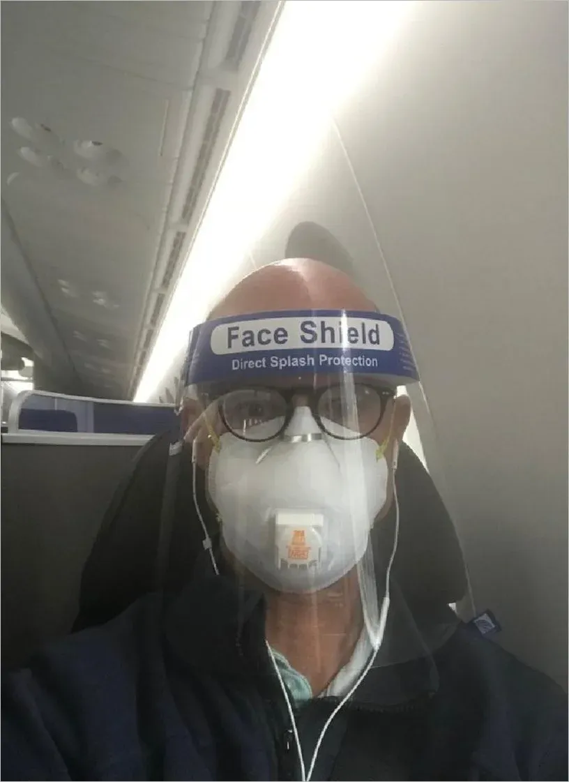 Selfie of man on an airplane wearing both a face shield labelled Direct Splash Protection and a valved N95 respirator underneath, plus glasses