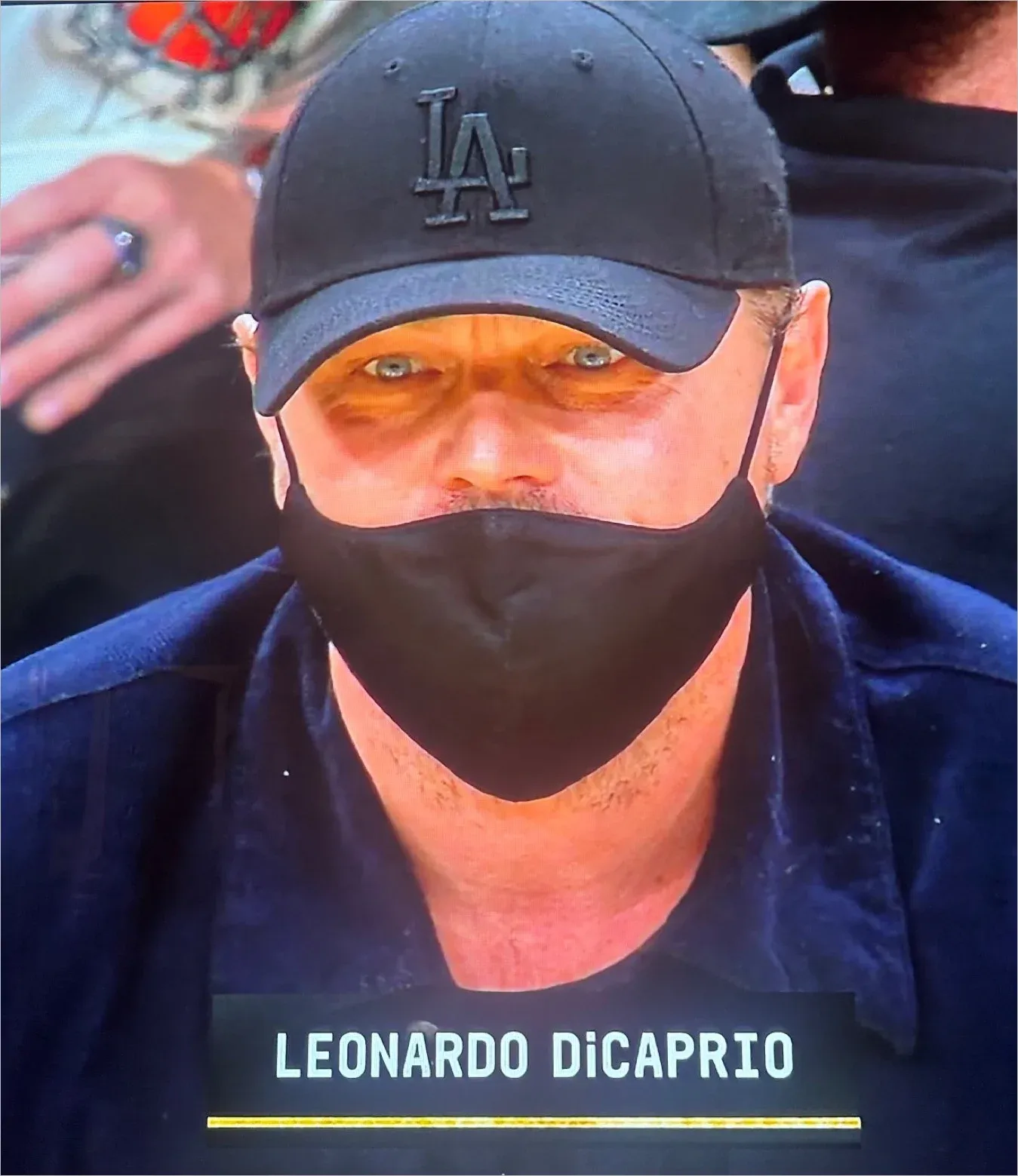 TV broadcast close-up of Leonardo DiCaprio at a sporting event wearing black face mask and LA baseball cap, name displayed on screen
