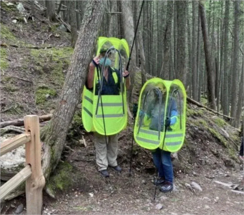 Two hikers on a forest trail wearing full-body yellow pop-up pod enclosures with clear plastic windows and reflective strips, plus masks underneath