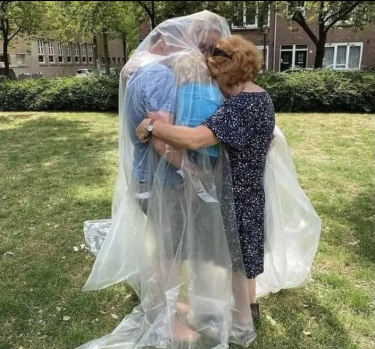 Photo of two people hugging outdoors with one person wrapped entirely in clear plastic sheeting to enable safe physical contact during COVID restrictions