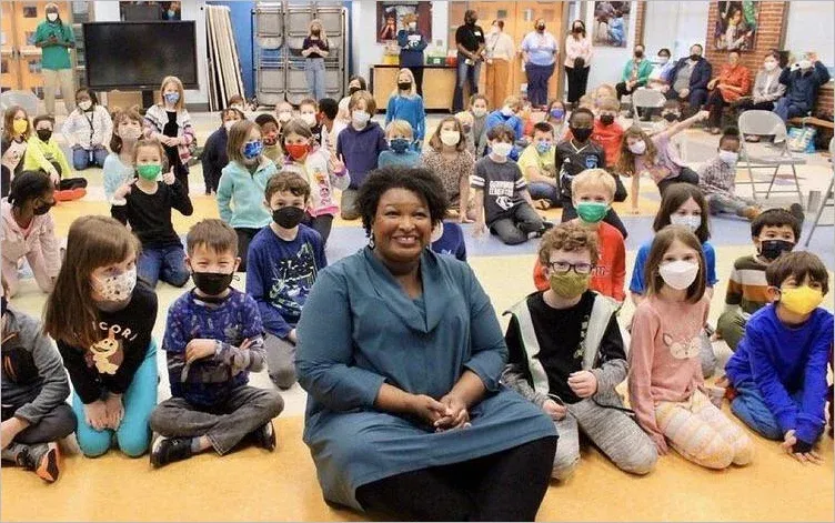 Photo of Stacey Abrams sitting unmasked and smiling among dozens of masked schoolchildren in a school hall