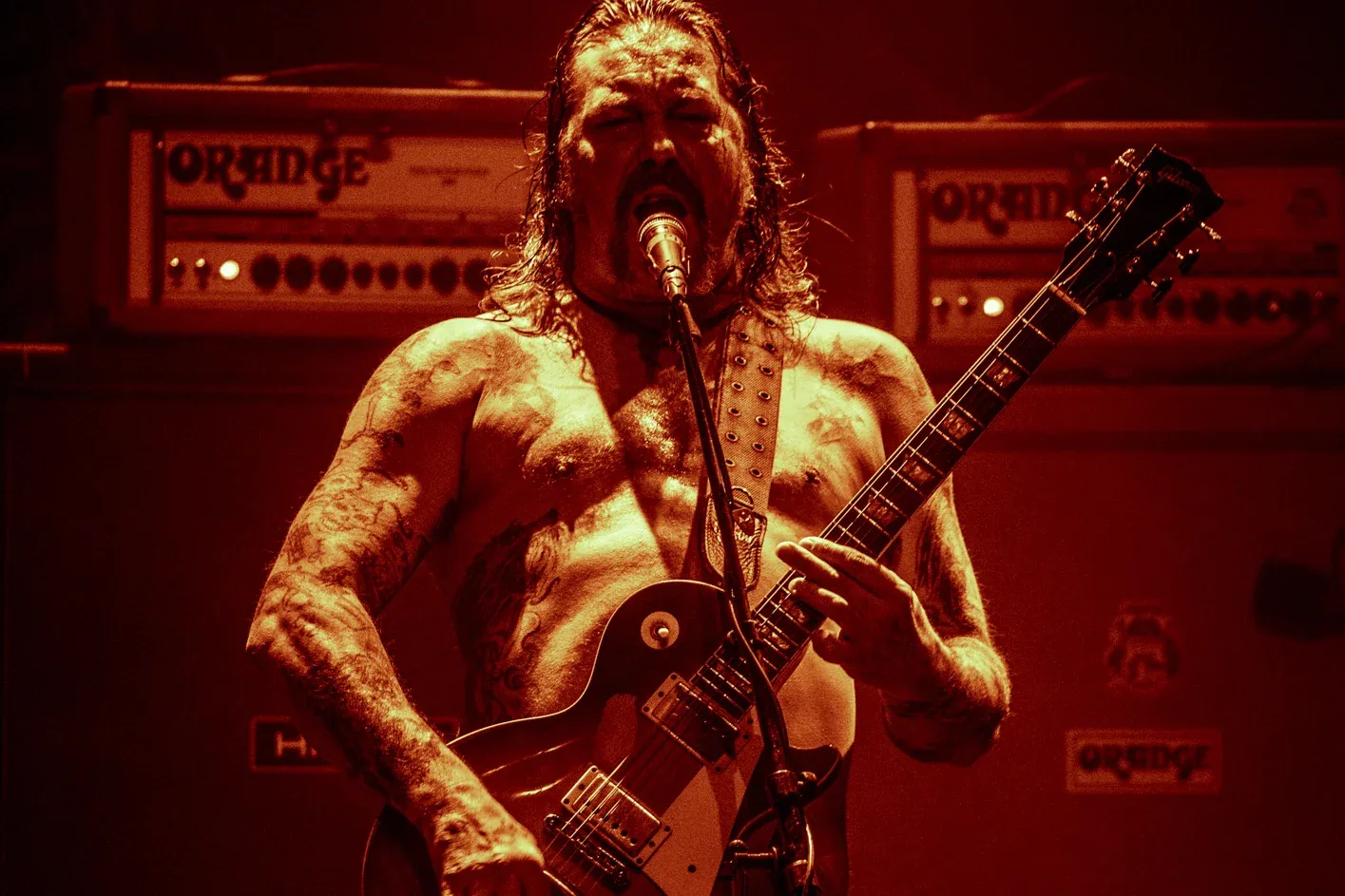 Matt Pike performing shirtless on stage, heavily tattooed, playing a Les Paul guitar bathed in red light with Orange amplifiers behind him