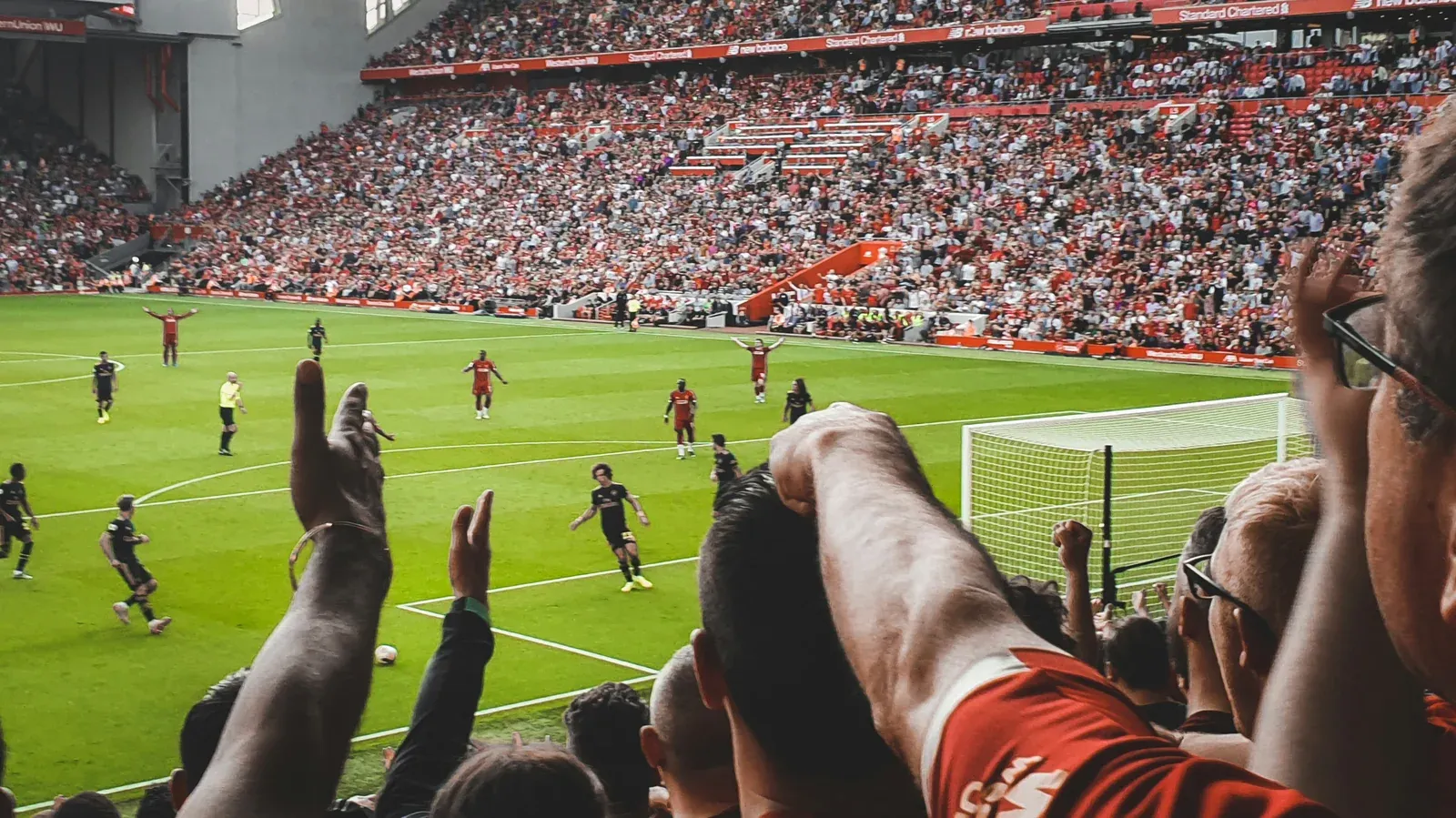 Football fans celebrating in the stands with fists raised as a match unfolds on the pitch below