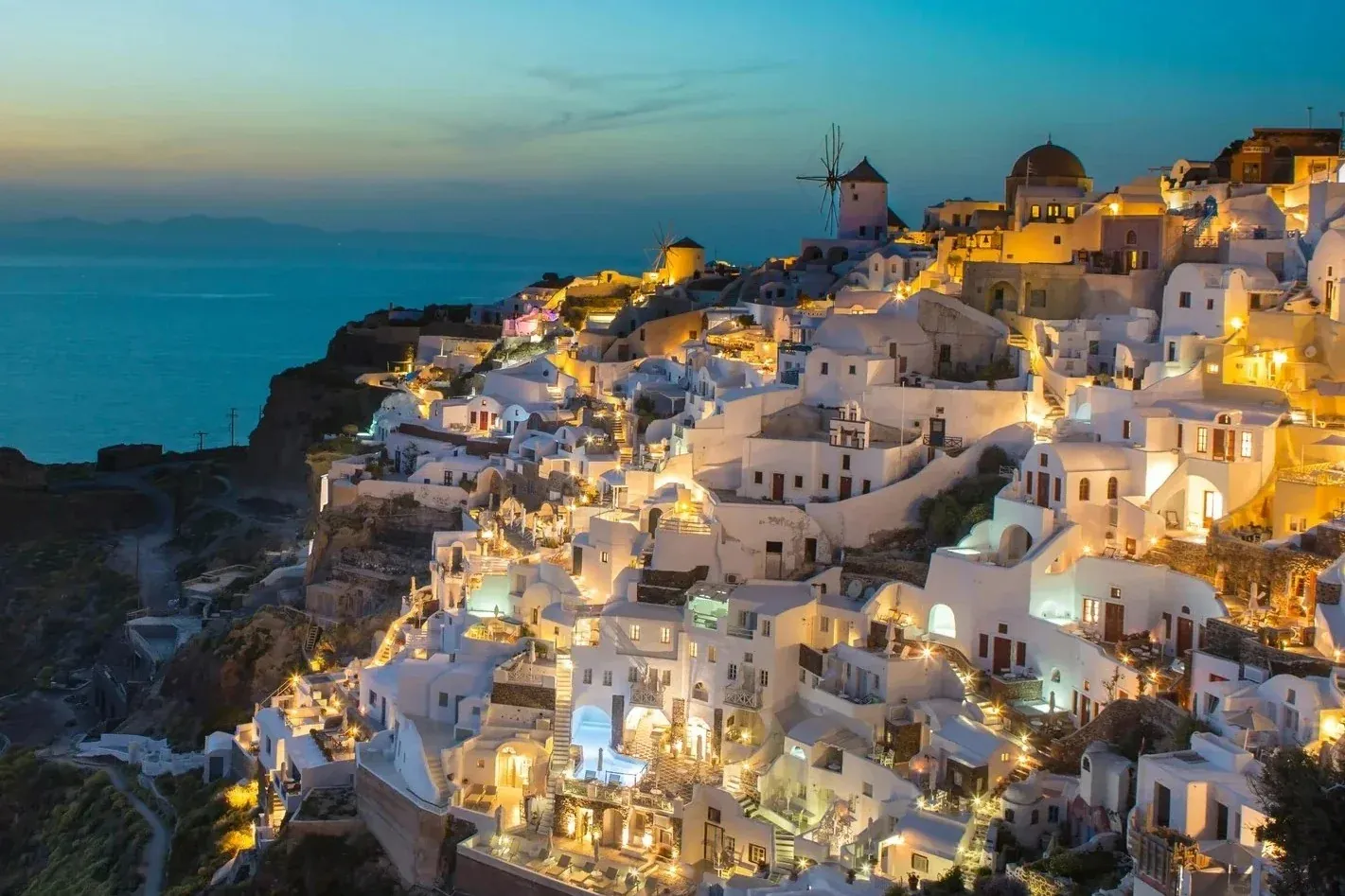 The white-washed buildings and windmills of Oia, Santorini lit up at dusk against the Aegean Sea