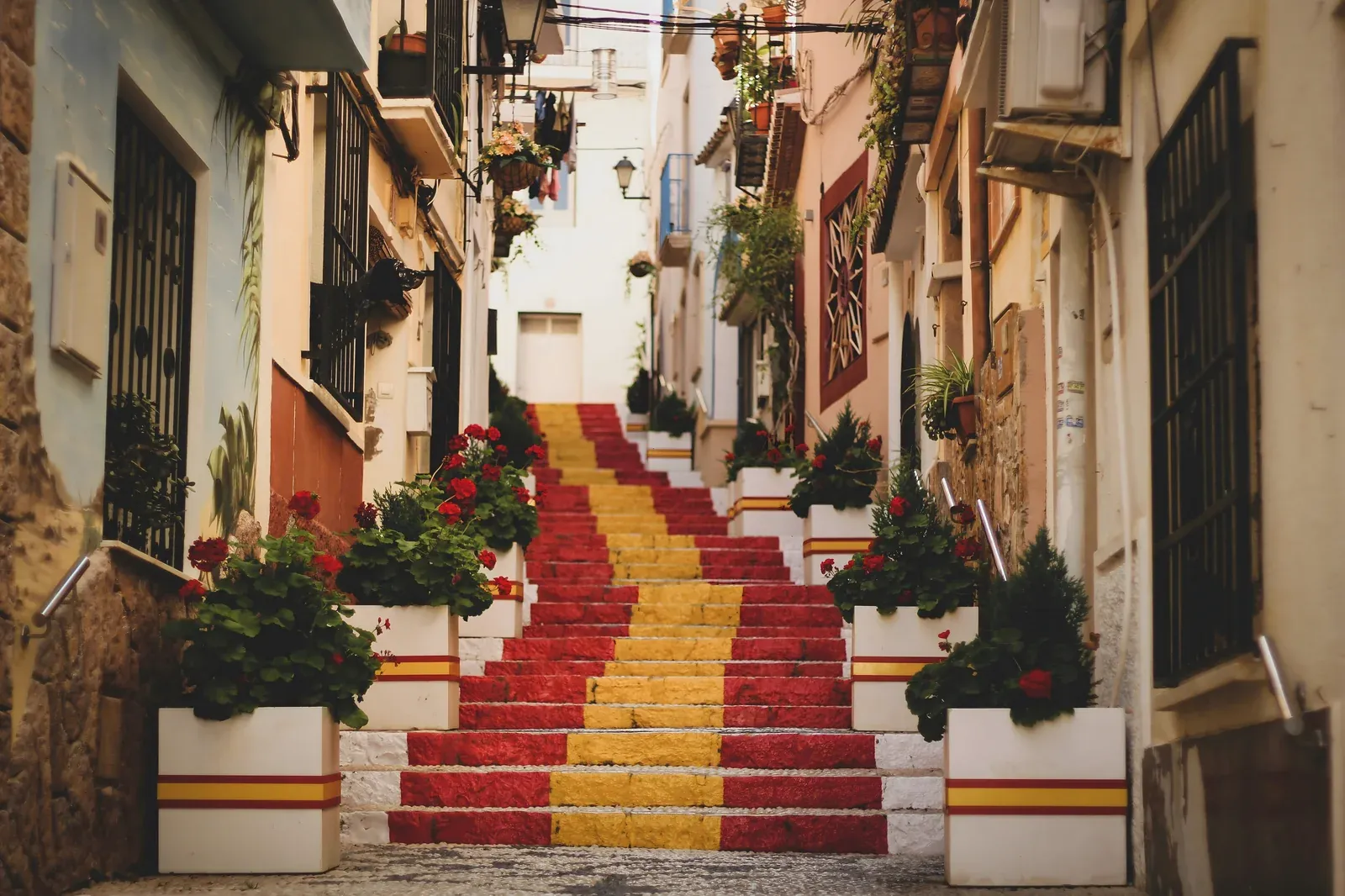 Steps painted in the red and yellow of the Spanish flag in a narrow old-town alley in Calpe, Spain, lined with potted plants and flowers