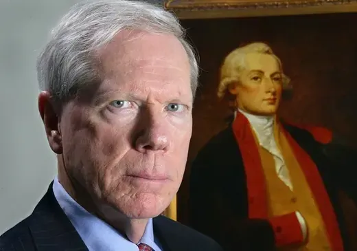 Paul Craig Roberts in a dark suit, looking directly at the camera with an oil painting portrait visible behind him