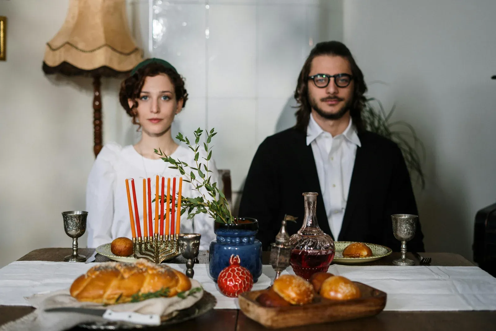 A young couple seated at a table set for a Jewish celebration with a menorah, challah bread, silver goblets and a decanter of red wine