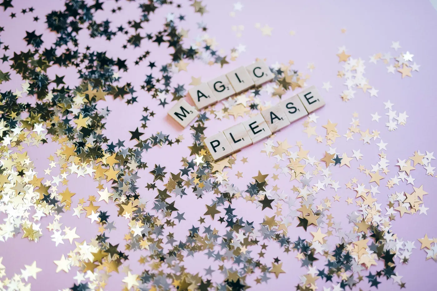 Scrabble tiles spelling 'MAGIC PLEASE' on a pink surface scattered with gold and silver star-shaped confetti