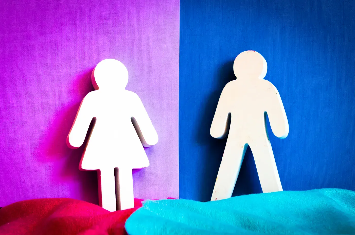 White wooden female and male figures against a split pink and blue background with red and turquoise fabric