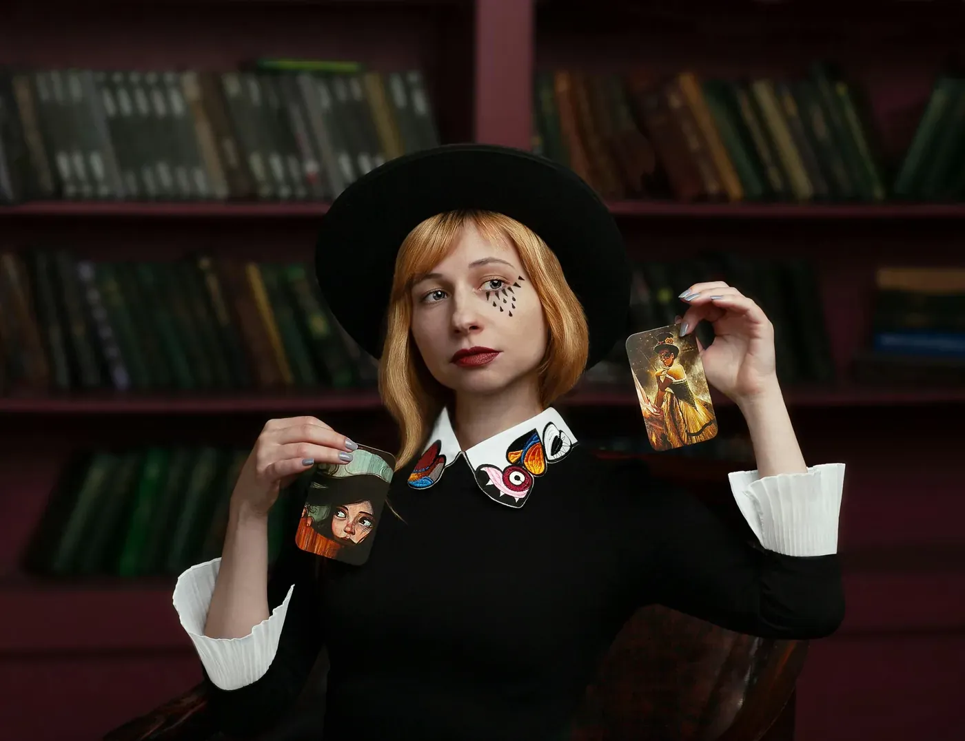 Red-haired woman in a black outfit and wide-brimmed hat holding up two illustrated cards in front of a library bookshelf