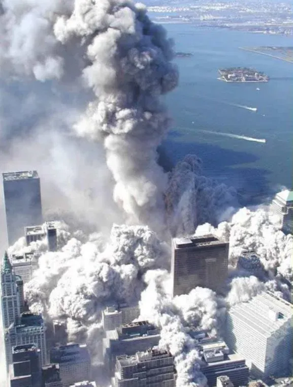 Aerial view of WTC collapse with dust cloud spreading outward at ground level while a tall column of debris rises vertically, harbor and islands visible in the background