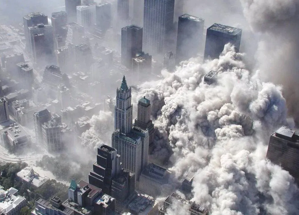 Aerial view of lower Manhattan with the Woolworth Building prominent in foreground, massive expanding dust cloud from WTC site behind it, surrounding area already coated white