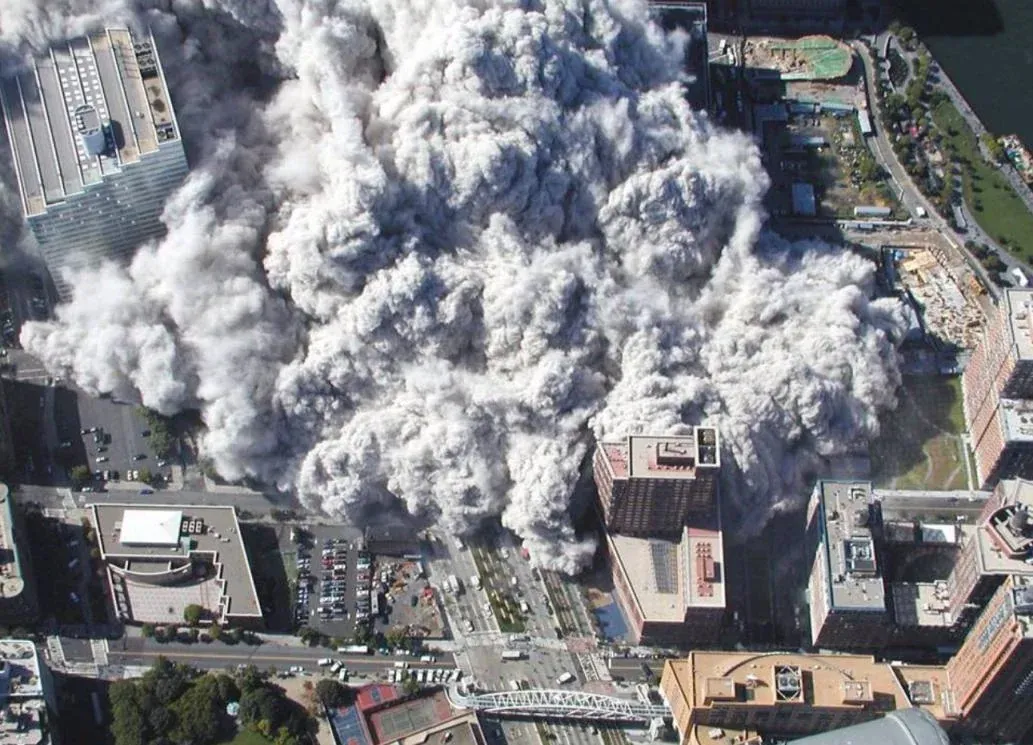 Aerial view from south looking down at WTC collapse, dense dust cloud engulfing nearby streets and buildings, Battery Park City and waterfront visible at top right
