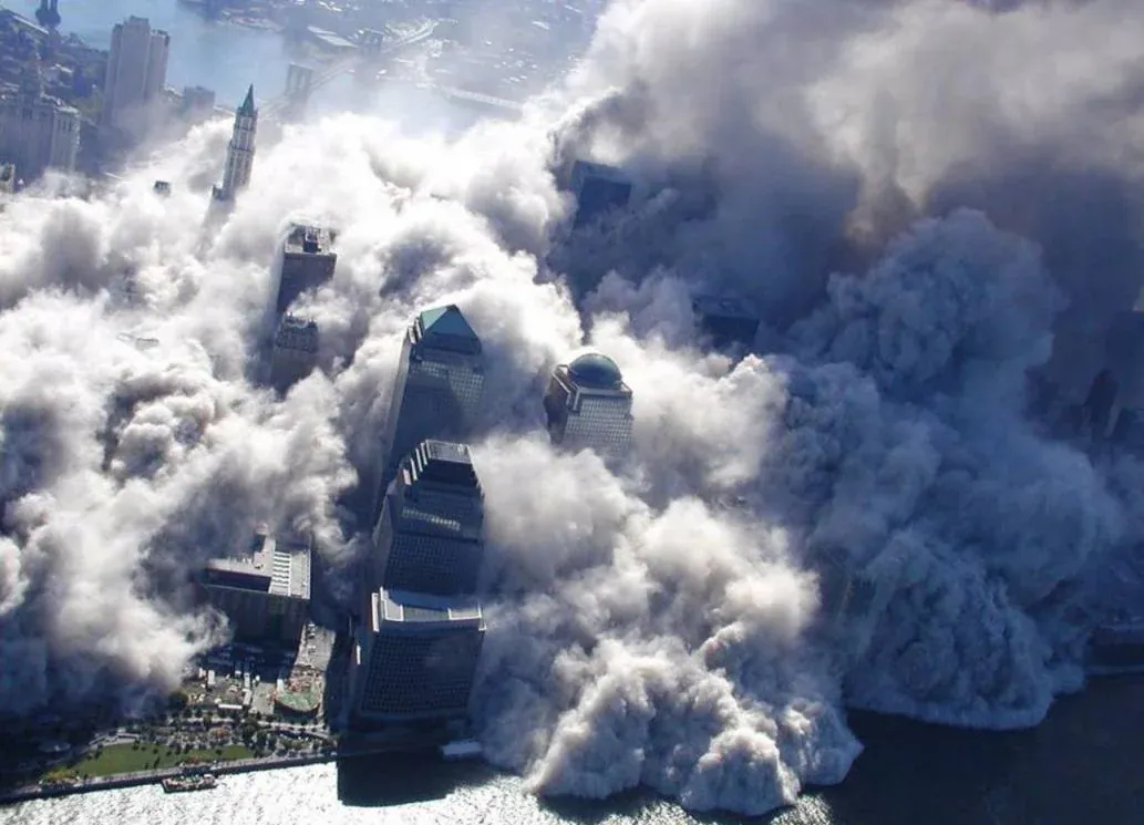 Aerial view from the west showing World Financial Center towers visible through a massive dust cloud rolling toward the Hudson River waterfront, dark smoke on the right side