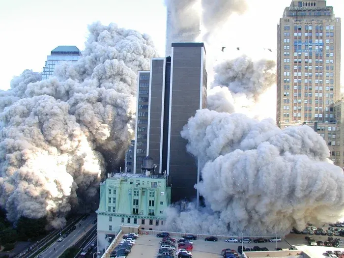 Street-level view of billowing dust cloud engulfing lower Manhattan buildings, debris visible in the air, green-roofed building in foreground, massive clouds surrounding high-rises