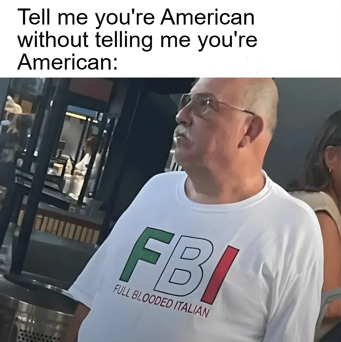 Meme photo of an older bald man with glasses sitting in what appears to be a restaurant, wearing a white t-shirt that reads "FBI - FULL BLOODED ITALIAN" in Italian flag colours (green, white, red). Caption at top: "Tell me you're American without telling me you're American:"