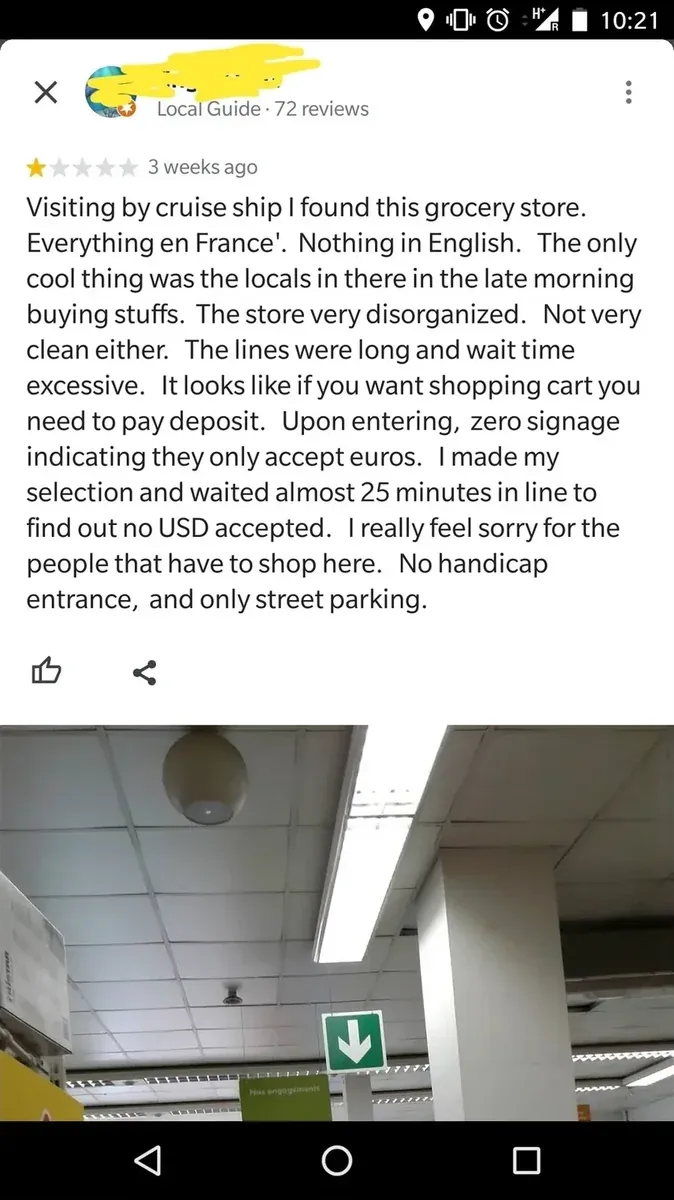 Google Maps review on mobile screenshot. Local Guide with 72 reviews (username redacted, 1 star, 3 weeks ago). Text: "Visiting by cruise ship I found this grocery store. Everything en France'. Nothing in English. The only cool thing was the locals in there in the late morning buying stuffs. The store very disorganized. Not very clean either. The lines were long and wait time excessive. It looks like if you want shopping cart you need to pay deposit. Upon entering, zero signage indicating they only accept euros. I made my selection and waited almost 25 minutes in line to find out no USD accepted. I really feel sorry for the people that have to shop here. No handicap entrance, and only street parking." Photo below shows store interior with ceiling and green exit sign
