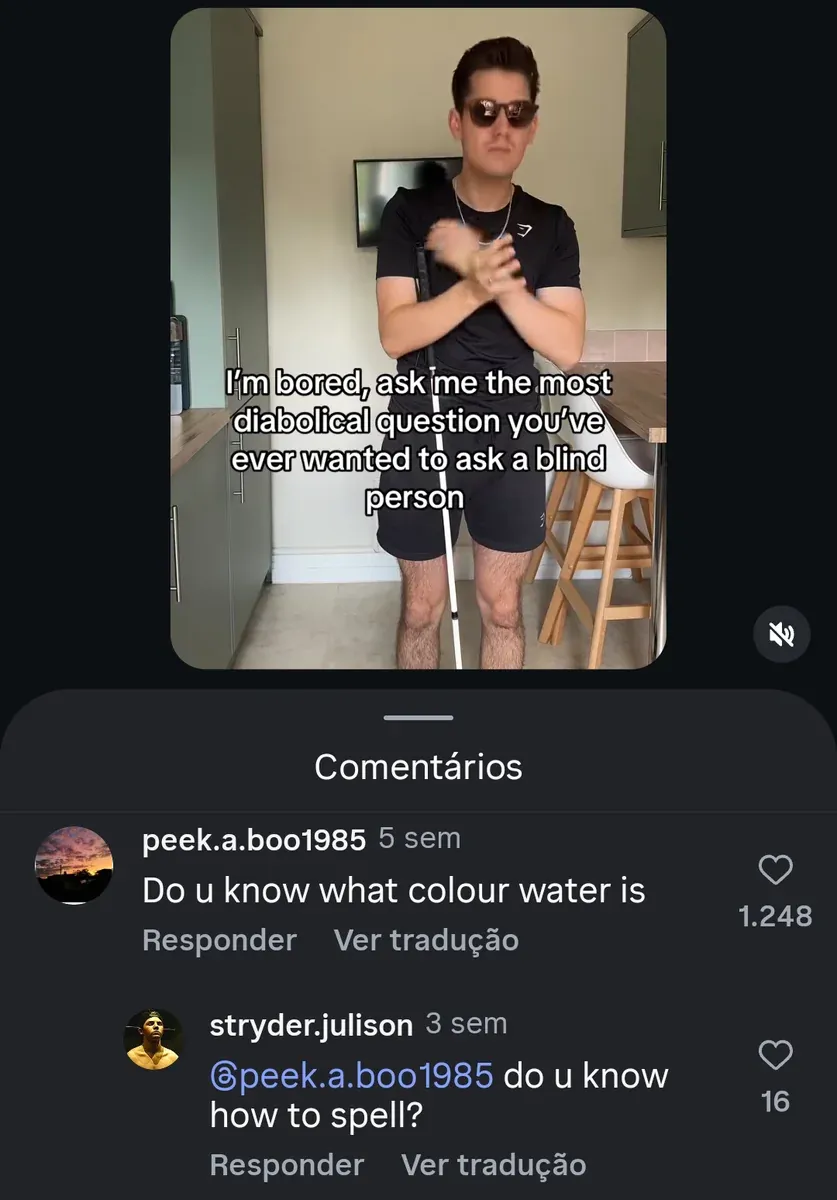 TikTok/Instagram post with Portuguese-language interface ("Comentarios", "Responder", "Ver traducao"). Video showing young man in black t-shirt and shorts standing in kitchen with sunglasses and a white cane (blind person), hands clasped. Caption on video: "I'm bored, ask me the most diabolical question you've ever wanted to ask a blind person". Comments: peek.a.boo1985 (sunset avatar, 5 sem): "Do u know what colour water is" Reply from stryder.julison (golden statue avatar, 3 sem): "@peek.a.boo1985 do u know how to spell?"